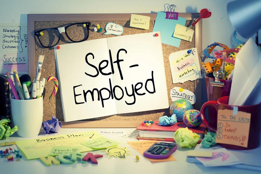 Picture of cork board on desk with book open with the words "self employed" written in it